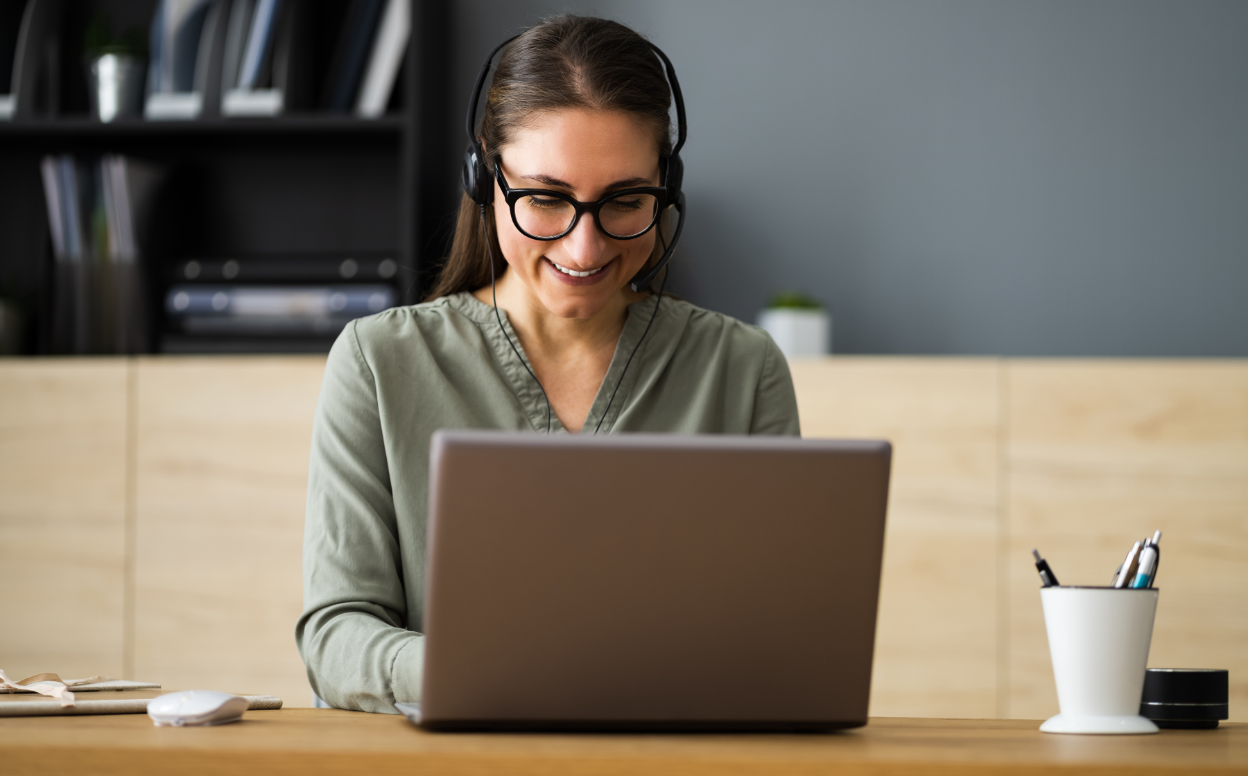 Woman wearing a headset and glasses smiling while working on a laptop