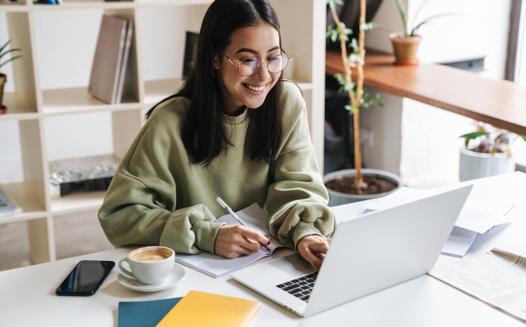 Young woman smiling while taking notes and working on a laptop
